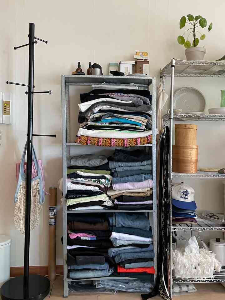 白い壁とモダンなスチール棚のある収納スペース、きちんと畳まれた衣類と屋内植物が配置された整理された空間