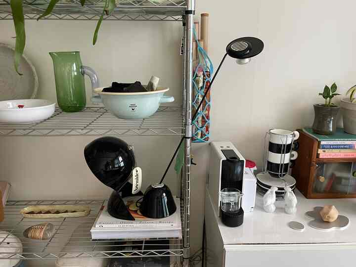 ホワイトの壁とテーブルにブラックの照明とコーヒーマシンが配置されたモダンなキッチンスペース、スチール棚に多様なオブジェが並ぶ