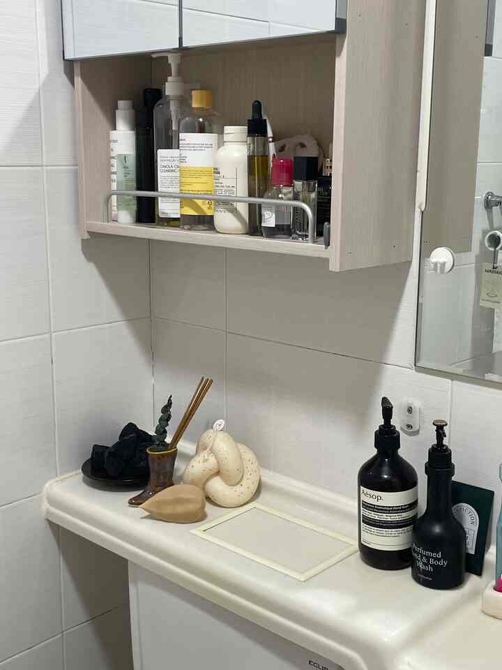 ベージュとホワイトの色調のバスルーム、上部に収納キャビネットがあり、洗面台の上にボディケア製品とお香が配置された雰囲気のあるインテリア