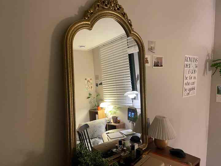 白い壁とゴールドのフレームが印象的な大きな鏡のある寝室、木目調ブラインドやダイニングチェア、テーブルランプが配置された雰囲気のあるインテリア