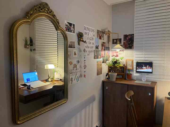 白い壁とブラインドを背景にしたヴィンテージの金縁壁鏡と木製の机が調和する居心地の良いホームオフィス空間