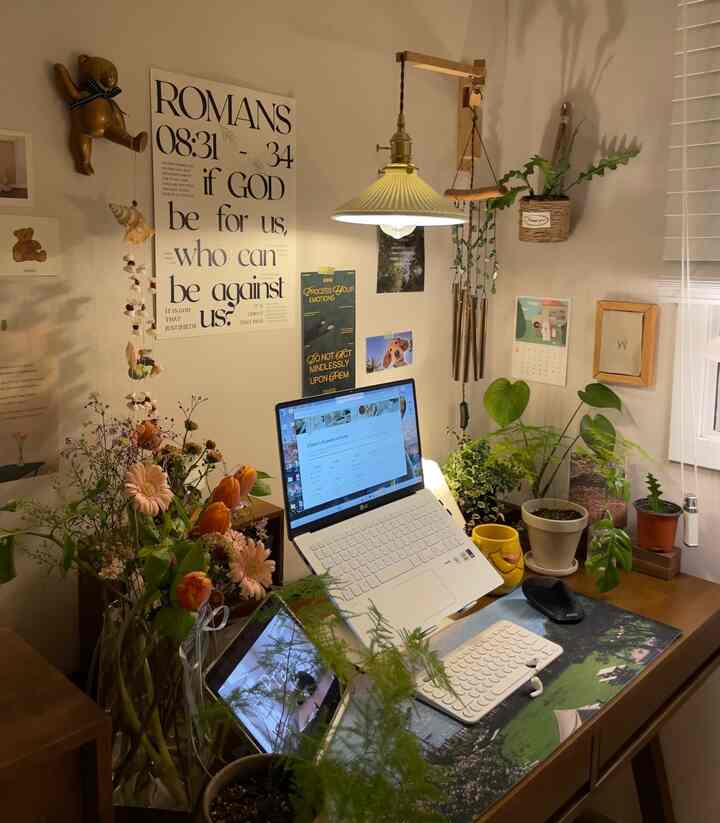 ナチュラルな色調のホームオフィス、木製のデスクに多数の植物が置かれ、ノートパソコンと温かみのあるペンダントライトがある雰囲気の良いインテリア