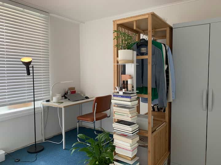 白い壁と青いカーペットのナチュラルなホームオフィス空間、木目調のオープンクローゼットとデスク、植物が配置された落ち着いた雰囲気のインテリア