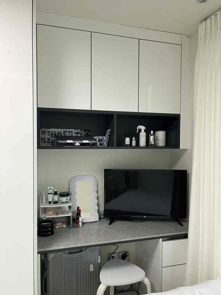 ホワイトトーンのコンパクトな寝室、化粧台兼用のデスクとテレビが配置された洗練されたモダンなインテリア