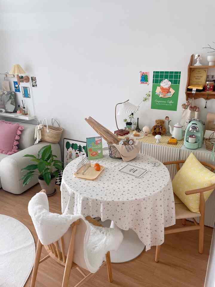 白い壁とベージュの木製床のナチュラルなダイニングルーム、花柄のテーブルクロスをかけた円形テーブルとホームカフェの要素があるインテリア
