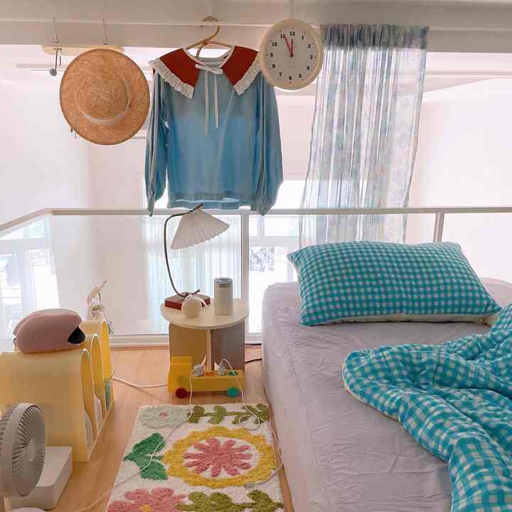 やわらかなブルーとホワイトのトーンが印象的なワンルームのロフト寝室、青いチェック柄の寝具とサイドテーブル、フロアランプが配置された居心地の良い空間