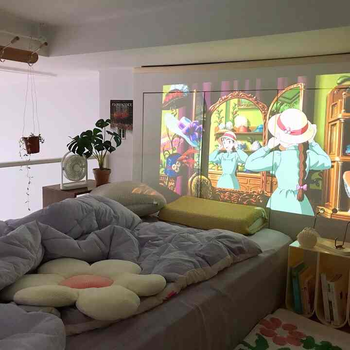 白い壁と天井が広がるロフト寝室、中央にベッドと花形クッションがあり、壁にアニメのプロジェクター画面が映し出された雰囲気のあるインテリア