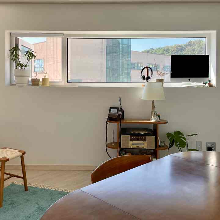 ナチュラルな色調の縦長の部屋、長い窓の下にデスクとダイニングテーブルが配置された明るい雰囲気の空間