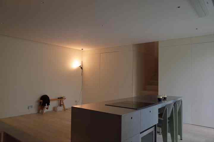 ベージュのトーンが広がるワンルームのキッチンスペース、ナチュラルな木のベンチとモダンなキッチンアイランドが配置されたシンプルで落ち着いた雰囲気のインテリア