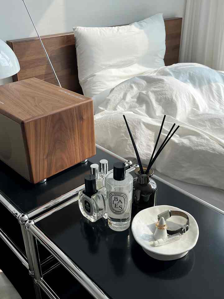 ウッドトーンのヘッドボードと白い寝具が配置されたシンプルモダンな寝室、ナイトテーブルにはスピーカーやディフューザー、香水が置かれた雰囲気のある空間