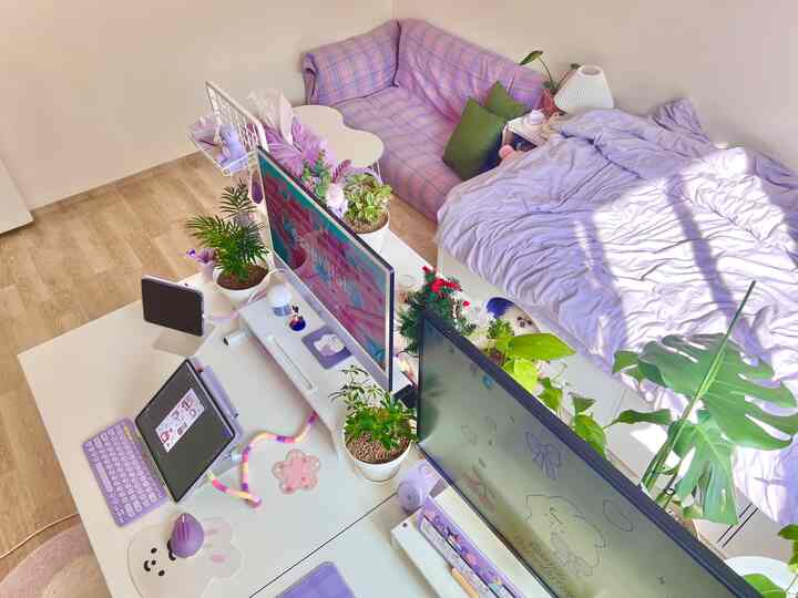 ラベンダー色調のワークスペース、デスクにデュアルモニターと植物が配置され、隣には寝具を敷いたソファベッドとベッドのある居心地の良い一人暮らしの部屋。