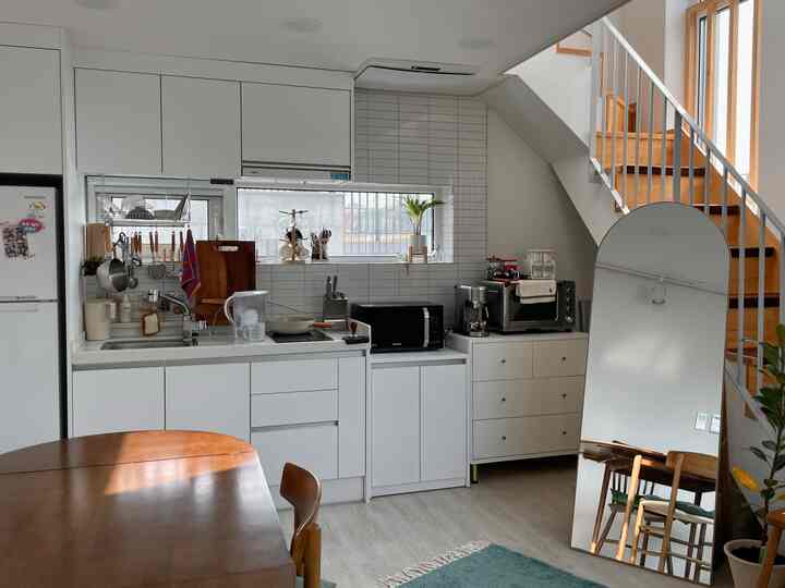 明るい白とナチュラルウッドのトーンのキッチンとダイニングテーブル、大きな鏡と階段が配置された居心地の良いワンルームスペース