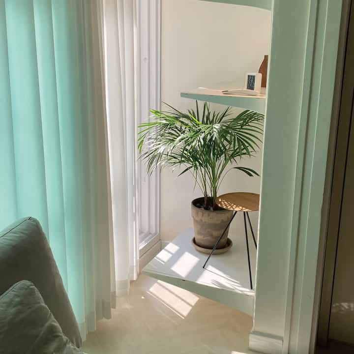 ホワイトとグリーンの色調の隙間収納スペース、壁の棚に鉢植えと窓辺のカーテンが配置された印象的な空間