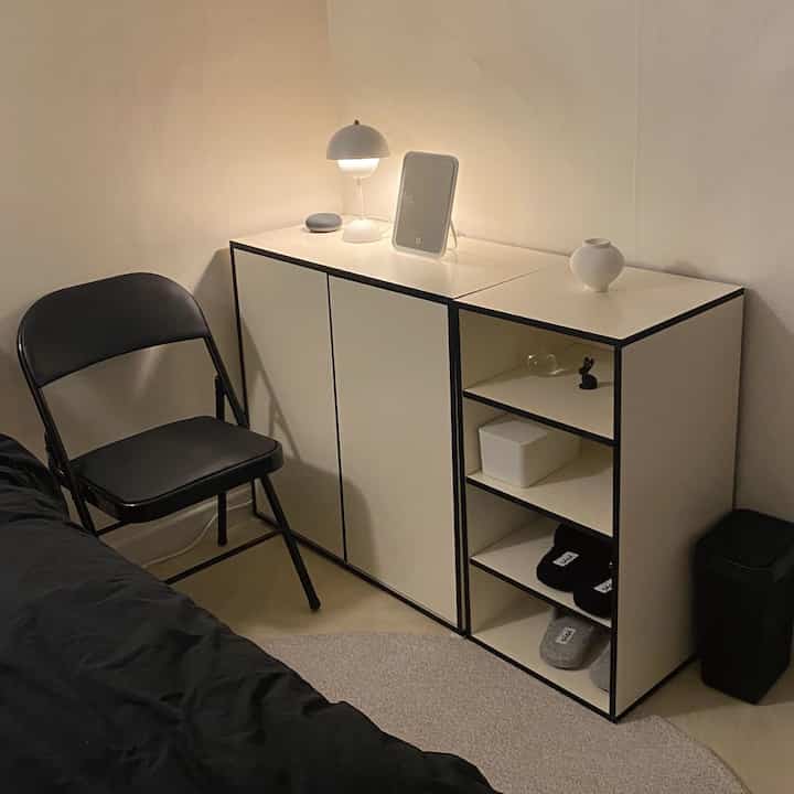 白い壁とモダンな収納キャビネットがある寝室の一角に、黒い寝具と椅子が配置されたシンプルな空間