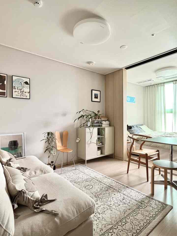 アイボリーと木目調が調和する1LDKリビング、シンプルなソファと額縁、ダイニングチェアが配置された落ち着いた空間