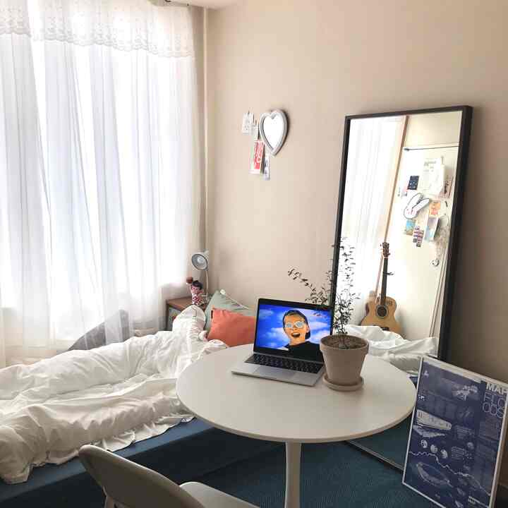 白いカーテンとベッド、丸いテーブルにノートパソコンと植物が配置された一人用シンプルモダンな寝室の空間