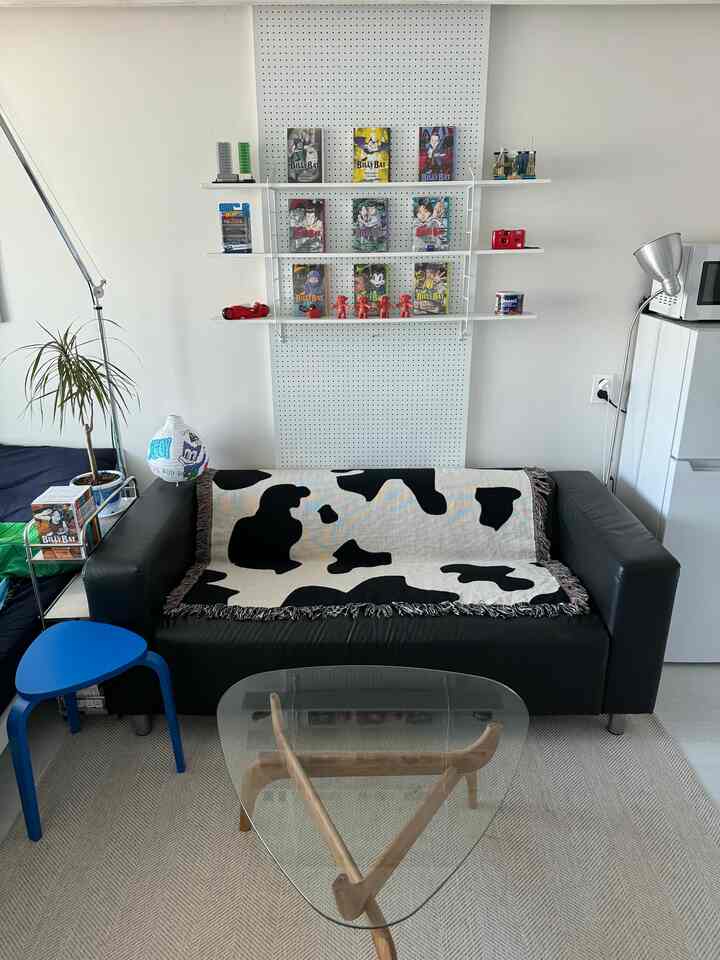 黒と白のトーンのリビングルーム、黒いレザーソファに牛柄のブランケットと壁の棚が配置された印象的な空間