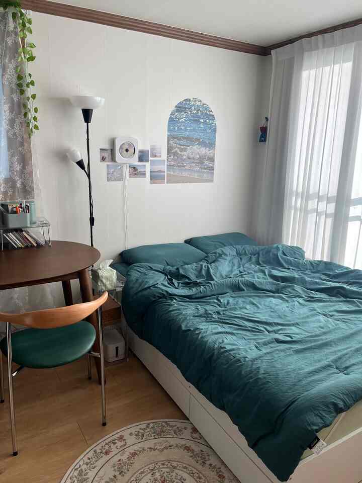 白い壁と青い寝具が印象的な寝室、丸い木製の机と透明なトロリー収納が実用的な雰囲気を演出している