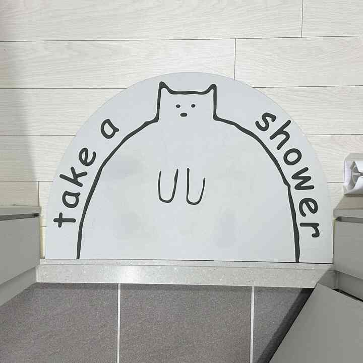 白色調のバスルーム入口の床に、かわいい猫のイラストが描かれた半円形の珪藻土バスマットが配置されている