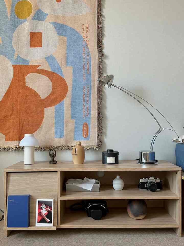 ベージュと木目調のリビング、壁掛けラグと小さな照明や小物が配置された温かみのある空間