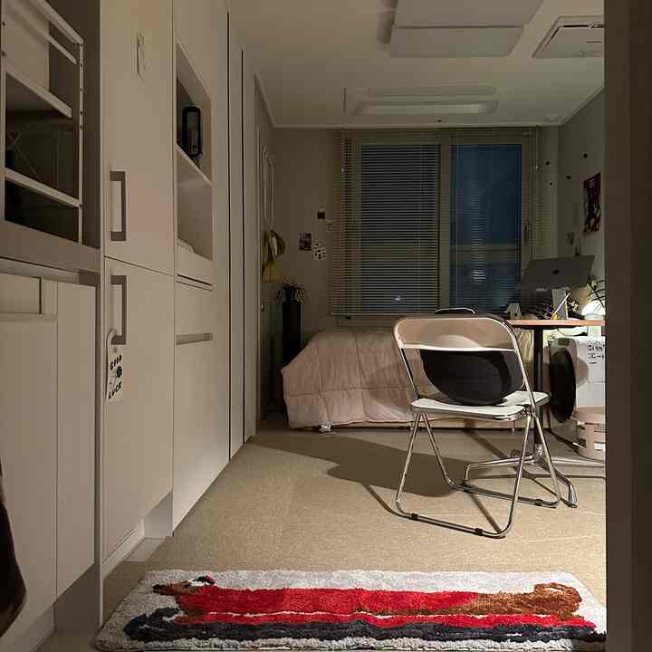 ベージュ色調のワンルーム、窓のブラインド、デスクと椅子、ベッド、床にカーペットと赤い模様のラグが配置された雰囲気のあるインテリア