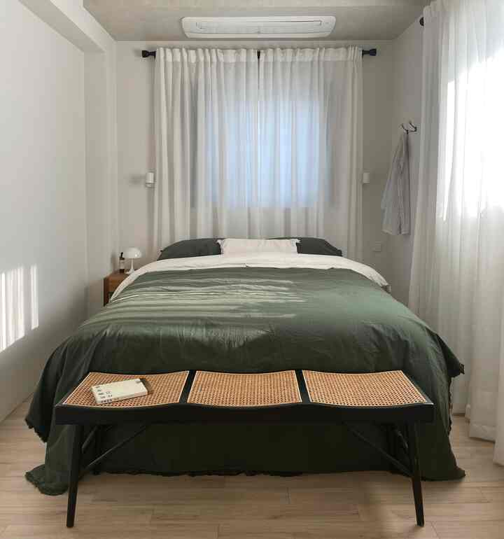 白いカーテンとナチュラルウッドの床が調和した、グリーンの寝具とベンチが配置された落ち着いた印象的な寝室