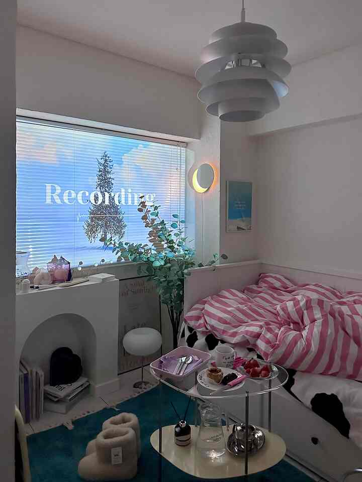 ピンクのストライプのベッドカバーがある小さな寝室、青緑のラグと窓際の植物が落ち着いた雰囲気を醸し出す