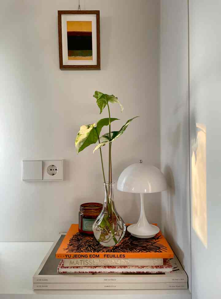 白い壁のコーナーにある小さな棚の上に植物とガラスの花瓶、テーブルランプ、キャンドルが置かれたミニマルな空間