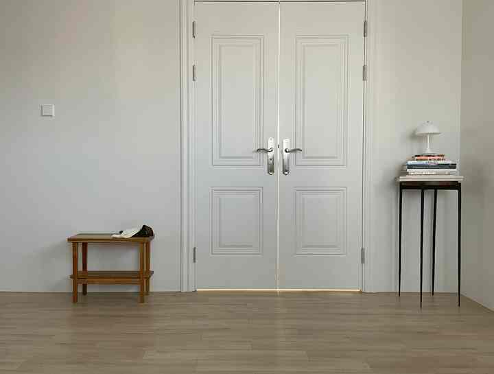 白い壁とドア、淡い木目調の床、左側にベンチ、右側にサイドテーブルとランプが配置されたシンプルなバスルームの入口空間