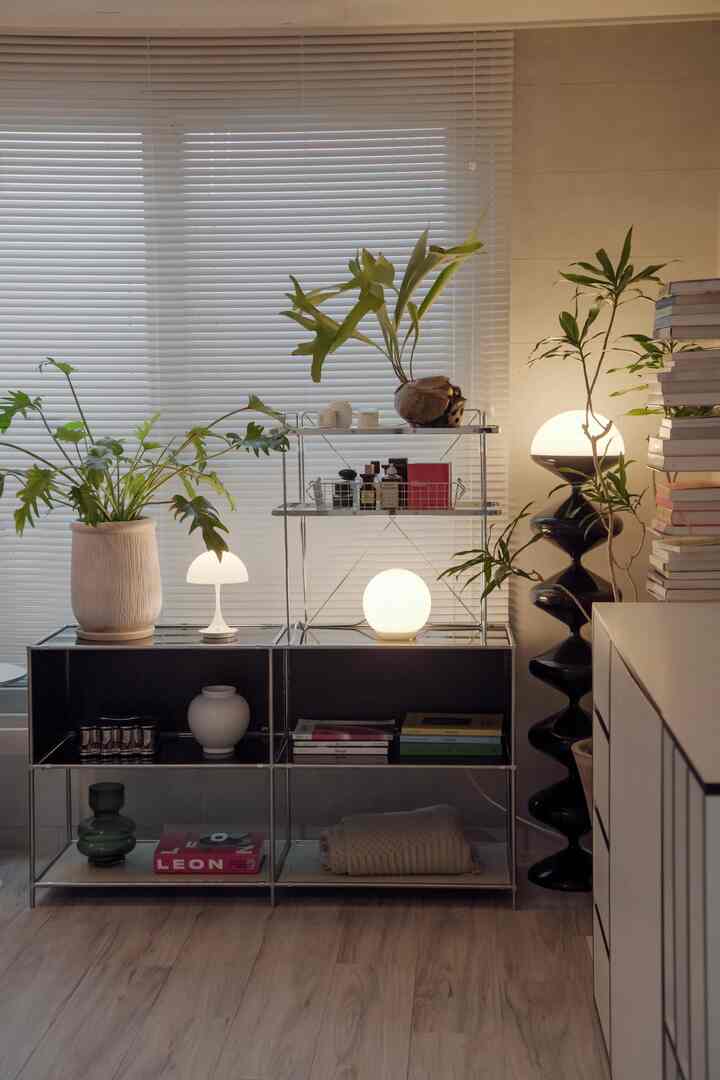 白いブラインドと木目調床のリビングスペース、モジュール棚と多様な植物や照明が配置されたミッドセンチュリーモダンスタイルのインテリア