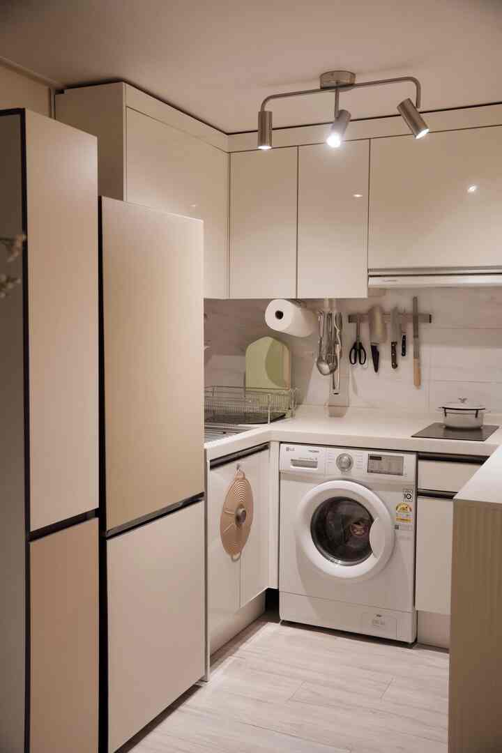 白とライトブラウンのトーンのモダンなキッチン、冷蔵庫と洗濯機、照明、食器乾燥ラックが配置されたすっきりとした空間
