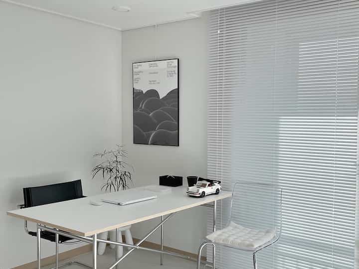 ホワイトトーンのホームオフィス、ホワイトテーブルとブラインド、黒いアームチェアが配置されたミニマルでモダンなインテリア