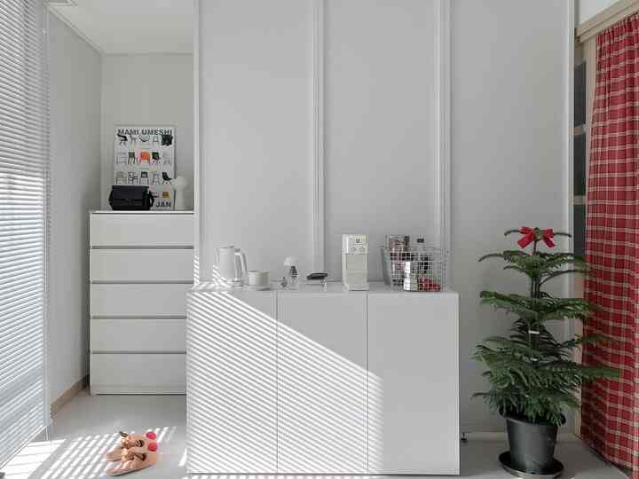 白を基調としたリビング、整頓されたキャビネットと左側のブラインド、右側にリボン付きアラウカリアの植物が配置された落ち着いた雰囲気のインテリア