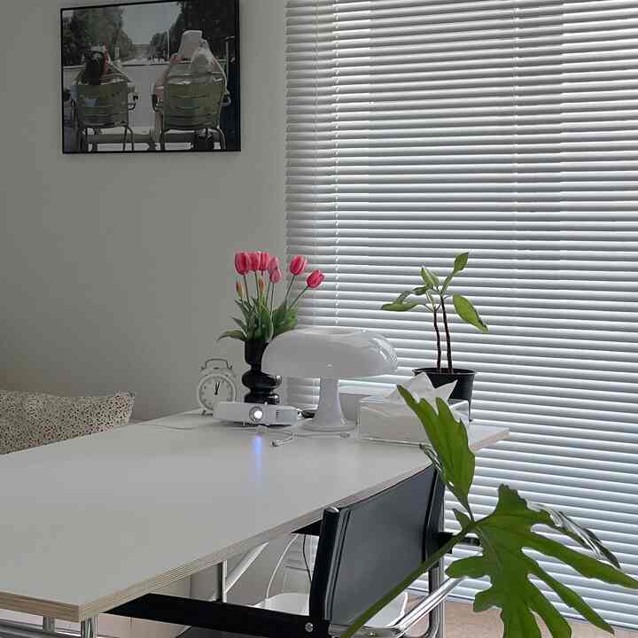 白色調のリビングまたは作業スペース、机の上にテーブルランプと植物、白いブラインドが配置された清潔感のあるモダンなインテリア