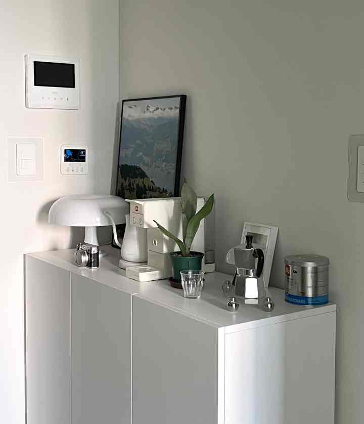 ホワイトトーンのキッチン、清潔なキャビネットの上にコーヒーマシンと植物、ポスターが配置されたモダンでシンプルなおうちカフェインテリア