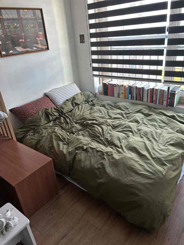 緑の寝具と木目調の床が特徴の6畳寝室、窓際の本とストライプのロールブラインドが配置された温かみのある空間