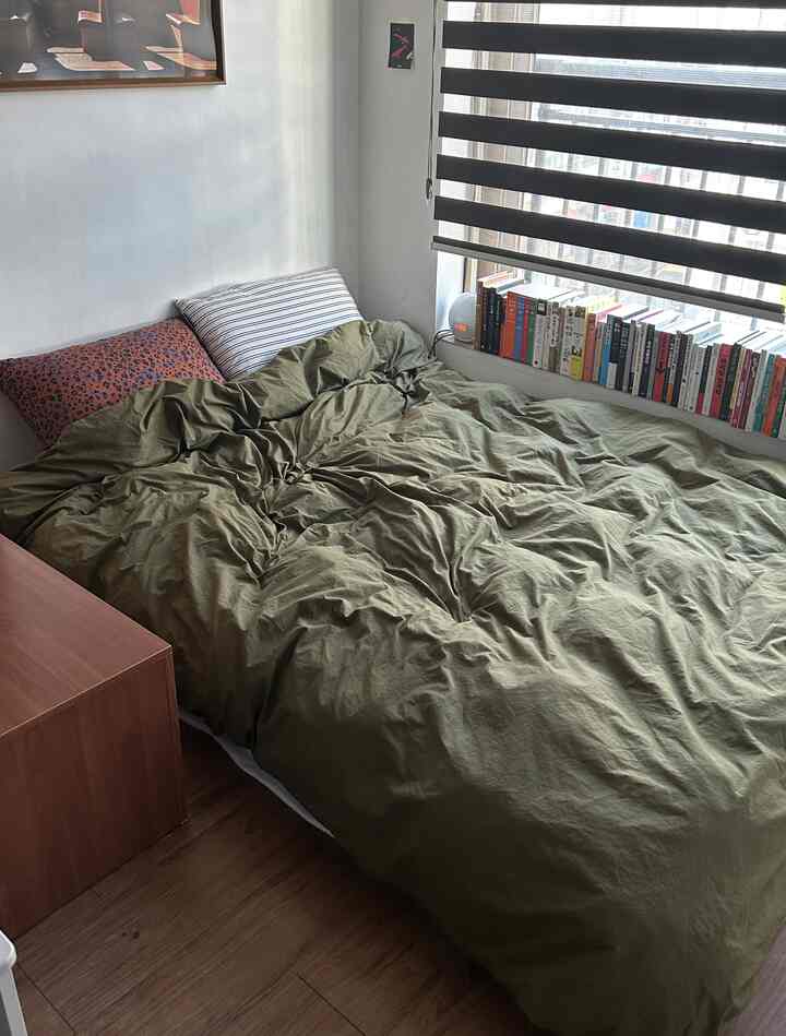 オリーブグリーンの寝具とブラウンの木製フロア、サイドテーブルが配置された落ち着いた寝室のインテリア