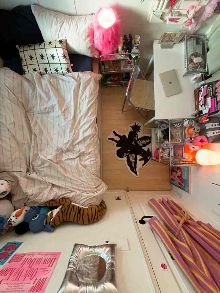 ピンク色の照明が映えるコンパクトな一人用寝室、白い机と折りたたみスツール、ぬいぐるみがベッドの横に並び、壁にはポスターが飾られた雰囲気のあるインテリア