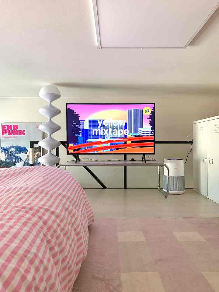 白とピンクの色調のリビング兼寝室スペース、中央にテレビ、左にフロアランプ、右に空気清浄機が配置された落ち着いたインテリア