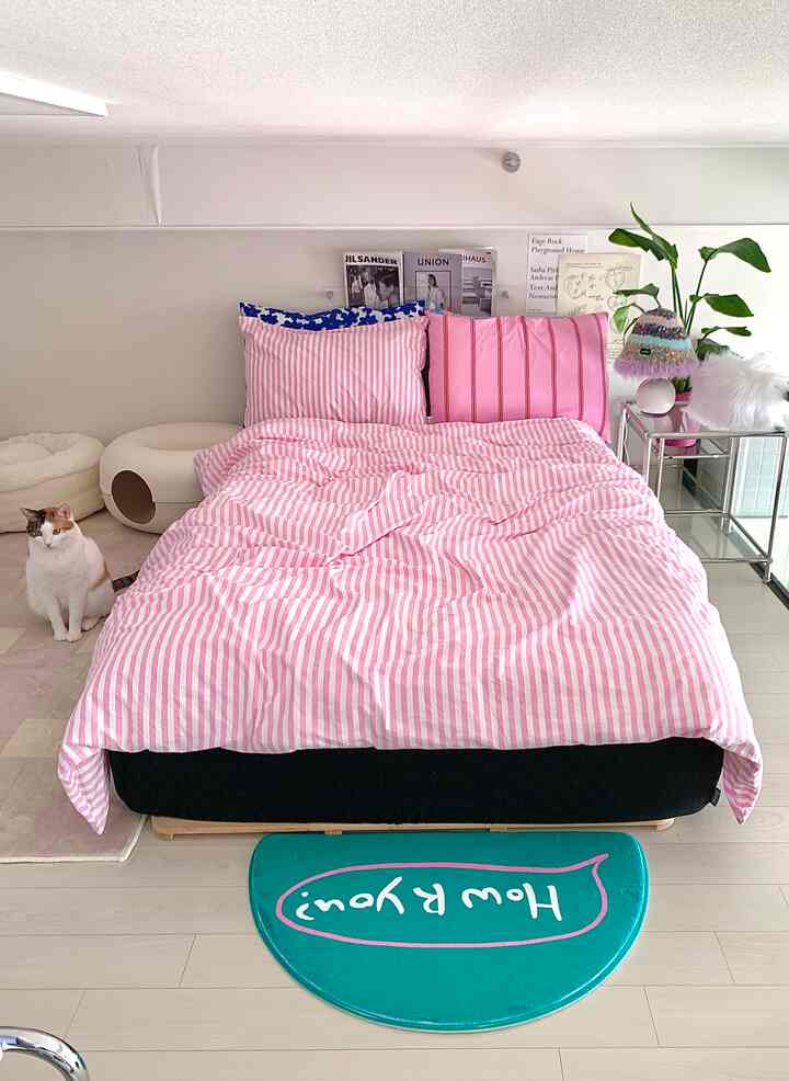 ピンクの寝具とラグが配置された寝室、黒いマットレスカバーがアクセントの居心地の良い空間