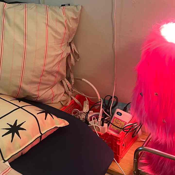 ネイビーの寝具と枕がある寝室のヘッドボード付近、赤い収納バスケットに充電器とマルチタップが整理され、ピンクのムードランプが空間を照らしている