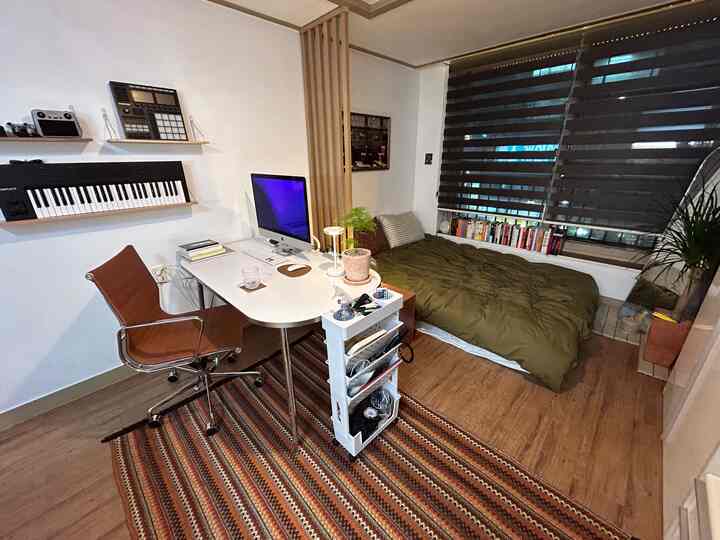 ナチュラルなブラウンウッドトーンとグリーンの寝具が調和する寝室兼ホームオフィス空間、スリムな木製間仕切りが配置された雰囲気のあるインテリア