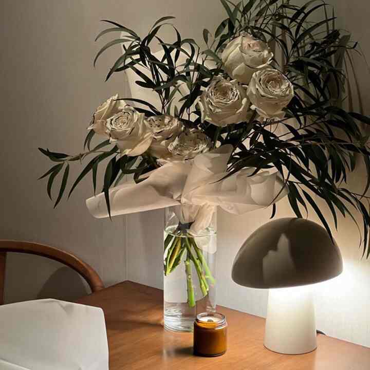 ナチュラルカラーとホワイトトーンが調和した寝室のテーブルに白いバラの花瓶と温かみのあるテーブルランプが置かれた雰囲気のあるインテリア