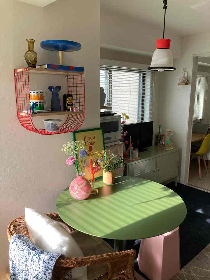 ナチュラルな色調の小さなダイニングルーム、緑の丸テーブルとラタンチェア、花の装飾と壁の棚が配置された雰囲気のある空間