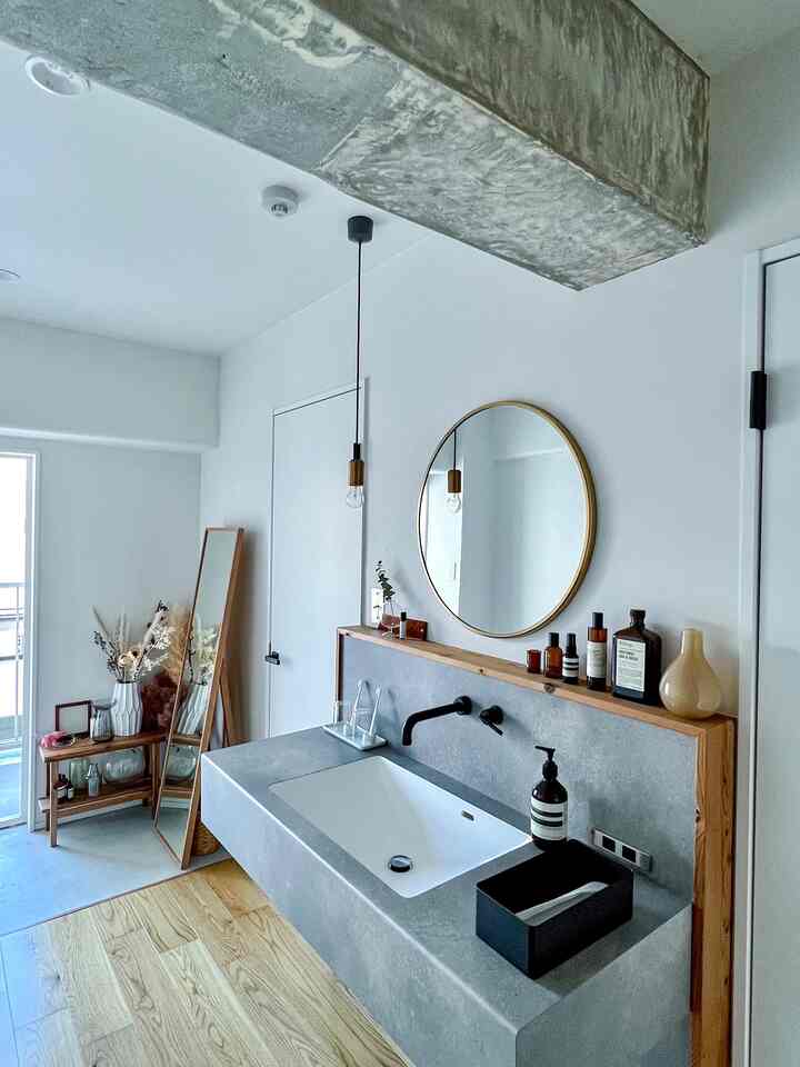 白い壁とグレーコンクリートの洗面台があるナチュラルな色調のバスルーム、丸い金縁の鏡とペンダントライトが配置された雰囲気のあるインテリア