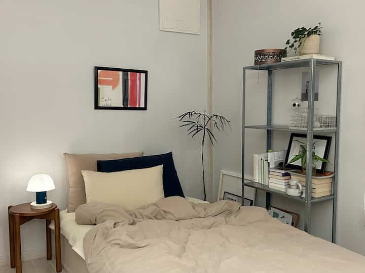 ベージュとネイビーカラーの寝室、ベッドと木製スツール、鉄製棚、テーブルランプが配置された落ち着いた雰囲気のインテリア