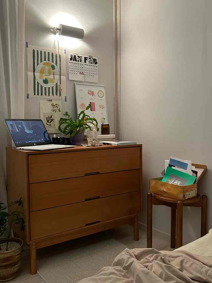 ナチュラルなトーンの寝室、木製の3段チェストと丸いスツール、壁に飾られた額縁アートが配置された居心地の良いホームオフィス環境