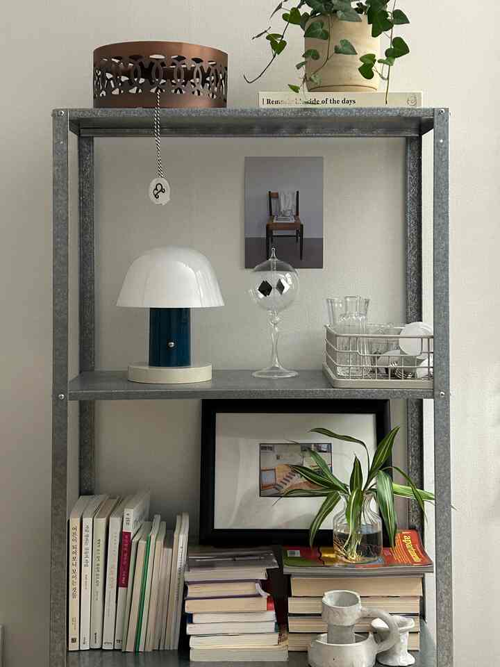 グレーのメタルシェルフに白いテーブルランプ、植物の鉢、額縁、整然と並んだ本が調和するモダンでナチュラルなインテリア空間