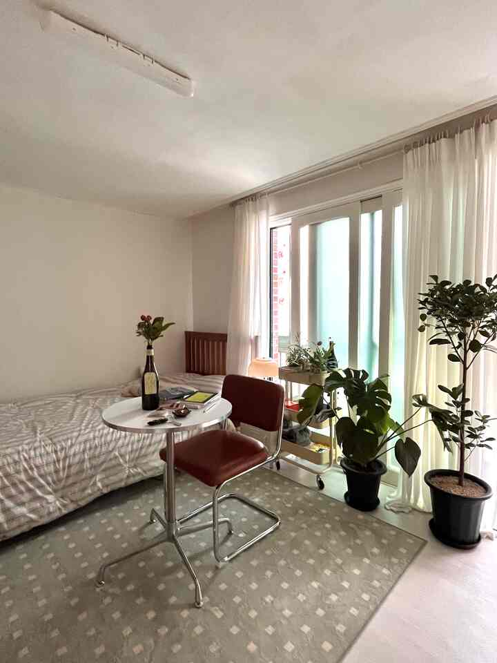 白とナチュラルトーンの縦長の寝室、ミッドセンチュリーモダンの椅子と丸テーブル、緑豊かな植物が配置された雰囲気のあるインテリア