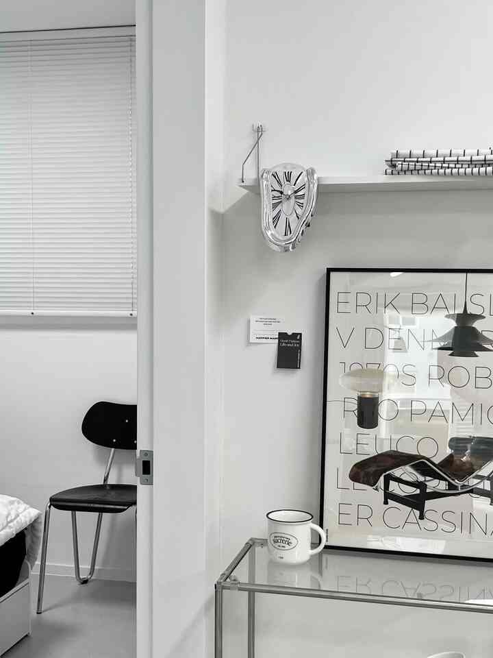 明るいホワイトトーンの寝室入口横のスペース、黒い椅子と壁の棚に置かれたユニークな時計、ガラステーブルとマグカップが配されたミニマルでモダンなインテリア